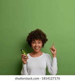 Vertical shot of glad woman with natural Afro hair holds bottle of freshly made smoothie, points above on empty space and wears casual white sweater. Healthy nutrtion concept. Lady holds detox drink - Powered by Shutterstock