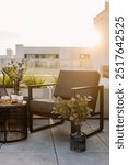 Vertical shot of gray chair near side table on balcony with cityscape view. Apartment with roof terrace, patio furniture, food, plants in vase and flowers. Warm sunlight creating a relaxing atmosphere