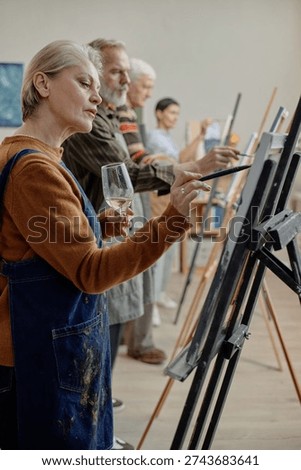 Vertical shot of group of middle aged and senior Caucasian adults painting on canvases in classroom setting, holding brushes and wine glasses, participating in educational art workshop