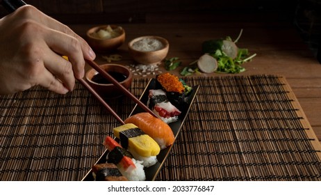 A vertical shot of hand picking up sushi from a plate of Japanese assorted sushi. Served with soya sauce on a black plate and with wooden background. - Powered by Shutterstock