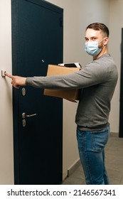 Vertical Shot Of Handsome Delivery Man In Medical Mask Ringing Doorbell Of Customer Apartment Looking At Camera, Holding Cardboard Box And Contactless Payment POS Wireless Terminal For Card Paying.