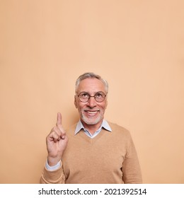 Vertical shot of happy bearded mature man points index finger above shows blank space for your text checks out advertisement big deal discount wears casual jumper isolated over beige background