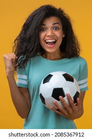 Vertical shot of happy football fan girl celebrating victory after betting at bookmaker website, making winner's gesture clenching her fist while holding ball in hand, isolated over yellow background.