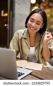 Vertical shot of happy girl talking on video call, looks at laptop, having online meeting, sitting in outdoor cafe. - Shutterstock ID 2233829795