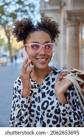 Vertical Shot Of Happy Pretty Millennial Girl With Two Hair Buns Calls Friend Via Smartphone After Making Shopping Carries Paper Bags Shares Impressions Wears Trendy Clothes Walks Outside Alone