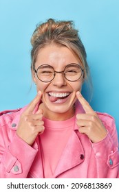 Vertical shot of happy woman points index fingers at cheeks sticks out tongue foolishes around feels glad wears round spectacles and pink jacket makes funny grimace isolated over blue background - Shutterstock ID 2096813869