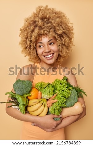 Vertical shot of happy woman poses with fresh green vegetables and fruits keeps to healthy diet wears casual t shirt isolated over brown background. Homegrown grocery. Vegeterian food concept