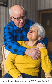 Vertical Shot Of A Hugging Elderly Caucasian Couple. Senior Gray-haired Woman Sitting On An Old-fashioned Chair Being Embraced By Her Bald Charming Husband In A Flannel Shirt. High Quality Photo