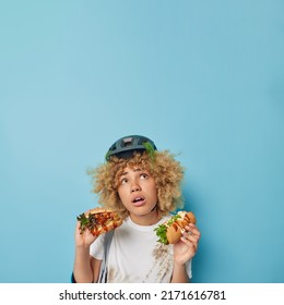 Vertical Shot Of Impressed Cury Haired Woman Wears Dirty T Shirt After Falling Off Bike Eats Fast Food Focused Overhead With Shocked Expression Holds Hot Dog And Hamburger Isolated On Blue Background