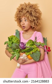 Vertical Shot Of Impressed Shocked Woman Carries Fresh Vegetables Fabric Bag Returns From Market Finds Out Impressive News Dressed In Pink Clothes Isolated Over Beige Background. Food Delivery