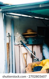 A vertical shot inside a storage area or shed, filled with assorted items and tools. Central elements include two straw hats stacked on a green crate, bags of supplies, and various gardening tools (sh
