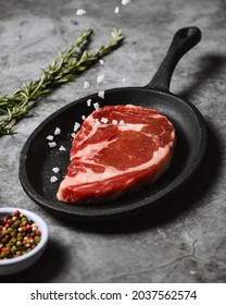 A Vertical Shot Of A Juicy Raw Steak On A Black Pan With Seasoning Falling On It On A Marble Table