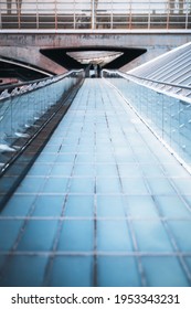 Vertical Shot Of A Long Modern Overhead Passage Made Of Blue Glass Tiled Blocks Stretching Into The Distance With Glass Fence And Chrome Banister Ending With A Pedestrian Tunnel Of A Railroad Station