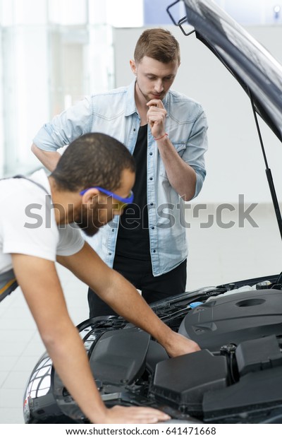Vertical shot of a male car owner looking\
thoughtful while professional mechanic is repairing his car at the\
workshop issues problems insurance checkup examination routine\
professionalism trust