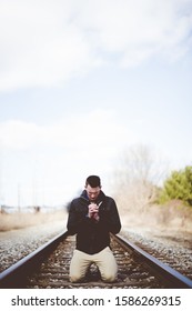 A vertical shot of a male on his knees on train tracks and praying while his eyes are closed
