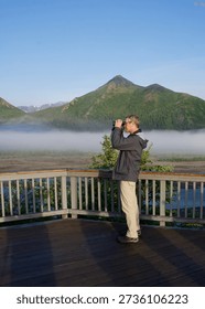 the vertical shot of a man using binoculars to look at the foggy mountains beyond the fence on a sunny day
