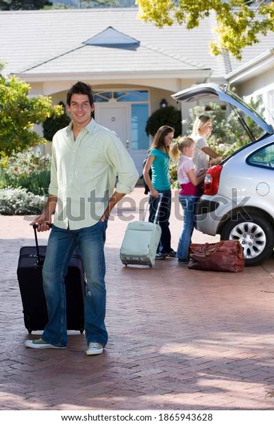 Vertical shot of a
mature man with luggage in tow with family loading luggage in car
boot in the
background.