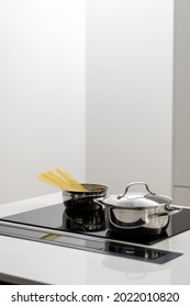 Vertical shot of metal pot with spaghetti and another one with lid closed on built in ceramic induction cooktop, food preparation in gloss kitchen counter with integrated cooking appliances