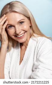 Vertical shot of middle aged woman in her 50s, has glowing natural face, nourished clear skin, smiling at camera, wearing bathrobe over blue background