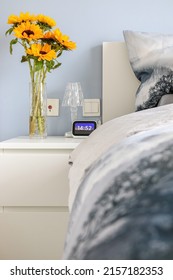 Vertical Shot Of Modern Elegant Bedroom With White, Blue And Grey Hues, Lit Up By Yellow Sunflowers (Helianthus) In A Glass Vase On Bedside Table, 14:52 On Digital Clock