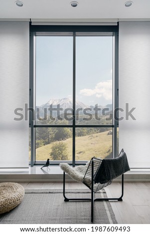 Vertical shot of modern metal chair on gray carpeted floor in front of large window with amazing mountain landscape in light coloured living room. Minimalist interior design in hotel room or house