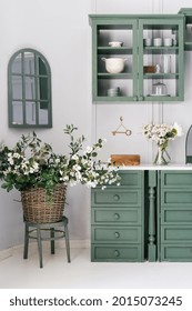 Vertical Shot Of Monochrome Kitchen Counter With Drawers And White Top, Hanging Cabinet With Glass Doors, Bowls And Mugs, Flowers In Jar And Woven Bucket, Vintage Furniture, Mirror With Window Shape