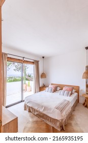 Vertical shot ofMediterranean villa guest bedroom in beige tones, wicker chandeliers, linens in local style and colors, a glass door leading to loggia overlooking the green rocky coastline