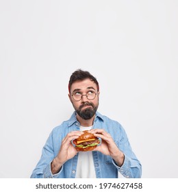 Vertical Shot Of Pensive Bearded Man Holds Appetizing Hamburger Focused Above Thinks Deeply About Something Wears Round Spectacles And Denim Shirt Blank Copy Space Overhead For Advertisement