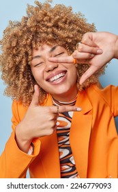 Vertical shot of positive curly haired woman smiles broadly keeps eyes closed makes frame with hands and fingers has happy face dressed in elegant orange jacket isolated over blue background