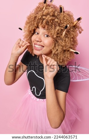Vertical shot of positive curly haired young woman cleans teeth with string takes care of mouth hygiene has blonde curly hair full of toothbrushes dressed like fairy isolated over pink background.