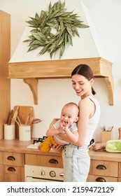 Vertical Shot. Positive Loving Caucasian Woman, Mother Playing With Baby At Home On Weekends, Mom Holding Son In Her Arms, Playing Together And Having Fun, Standing In Modern Kitchen.