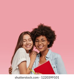 Vertical shot of positive mixed race young women or sisters have joy together, learn material, do home assigment, model against pink background with free space above for your advertising content - Powered by Shutterstock