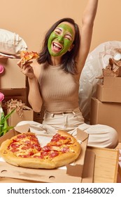 Vertical Shot Of Positive Young Asian Woman Undergoes Beauty Procedures Eats Tasty Pizza During Housewarming Party Keeps Arm Raised Surrounded By Cardboard Boxes Isolated Over Beige Background