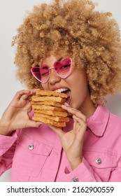 Vertical shot of pretty young woman with curly bushy hair bites big pile of waffles wears heart shaped sunglasses and jacket has sweet tooth poses indoor. Delicious appetizing homemade dessert