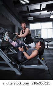 Vertical Shot Of A Professional Personal Trainer Assisting His Female Client In Leg Press Gym Machine Workout