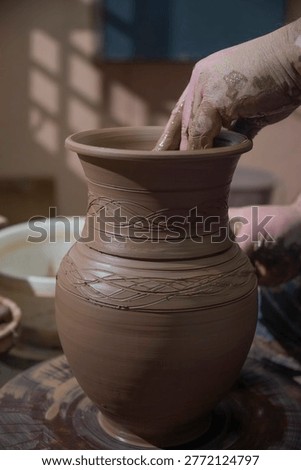 Vertical shot of a professional potter checking a tall clay vase with engraved ethnic patterns. Atmospheric workshop lighting with window shadows