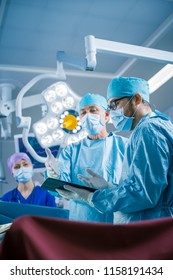 Vertical Shot Of A Professional Surgeon And Assistant Talk And Use Digital Tablet Computer During Surgery. They Work In The Modern Hospital Operating Room.