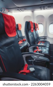 Vertical shot with selective focus on airplane seat in black fabric, seat belt, scarf over the headrest, and seat belt in bright red and in the background, more seats can be seen, and some windows