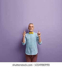 Vertical shot of self assured grey haired senior man points with both fore fingers upwards, dressed in denim shirt and yellow bowtie, spectacles isolated over lilac background with free space above