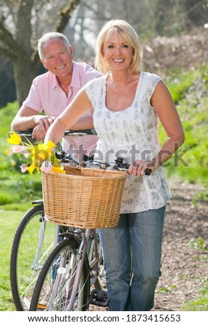 Foto vertical de una pareja mayor con sus bicicletas sonriendo en un día soleado en la cámara con la mujer delante llevando flores de primavera en su canasta.