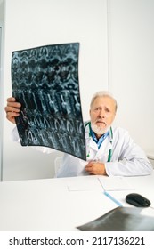 Vertical Shot Of Serious Mature Adult Male Doctor Holding MRI Spine Image Of Patient Sitting At Desk In Medical Office Room, Looking At Camera, Thinking About Diagnosis, Expressing Concern Of Illness.