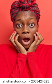 Vertical shot of shocked dark skinned young woman keeps hands on face stares bugged eyes mouth opened looks in stupefaction wears tied scarf on head casual jumper isolated over red background