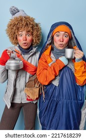 Vertical Shot Of Shocked Female Campers Stare With Omg Expression Drink Tea From Kettle Carry Wood Have Picnic In Nature Wear Winter Clothes Wrapped In Sleeping Bag Isolated Over Blue Background