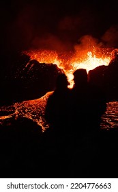 A Vertical Shot Of A Silhouette Of A Couple Watching Lava Flowing From A Volcano At Night In Iceland