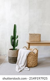 Vertical Shot Of Small Storage Box For Clothes On Wooden Bench Close To Wicker Basket With Cozy Plaid. Potted Cactus Near Coffee Table In Living Room, Standing Against Light Grey Wall With Copy Space