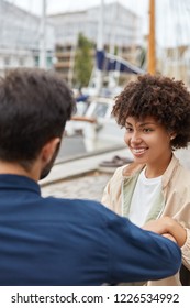 Vertical shot of smiling dark skinned girl has Afro haircut, toothy smile, enjoys friendly talk with Caucasian male friend, sit at bench near harbour, waits for someone. People, communication concept