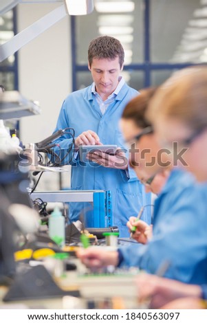 Vertical shot of supervisor using a digital tablet while watching technicians solder circuit boards on production line in a manufacturing plant.