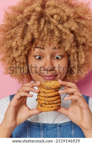 Vertical shot of surprised curly haired young pretty woman holds pile of appetizing chocolate chip cookies feels temptation to eat sweet food poses indoor. Yummy harmful food and calories concept.