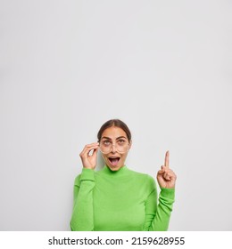 Vertical shot of surprised dark haired woman with amazed expression points index finger above demonstrates something unexpected dressed in green poloneck transparent eyeglasses poses indoor.
