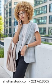 Vertical Shot Of Thoughtful Curly Haired Woman Has Telephone Conversation With Serious Expression Dressed Formally Carries Bag Stands Against Urban Background In Big City. Technology And Lifestyle